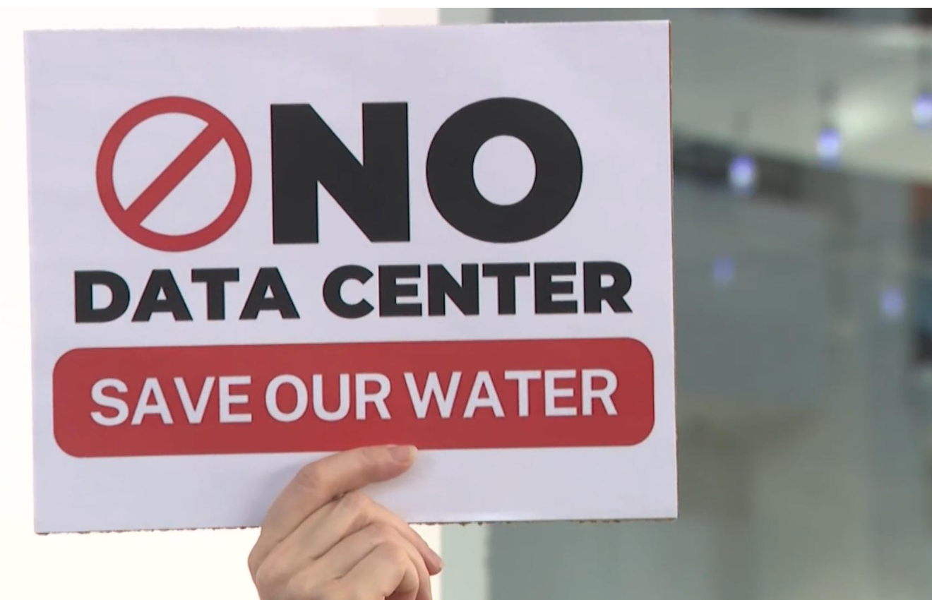 A hand holds up a sign reading "No Data Center — Save Our Water"